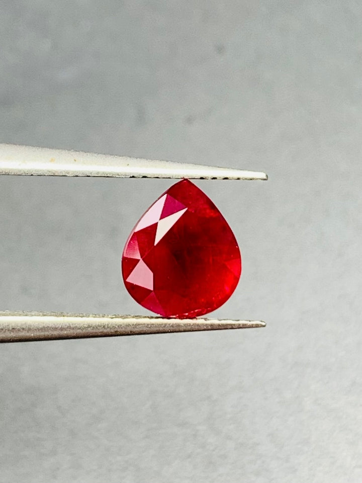 Teardrop-shaped Burma Ruby  gemstone held between tweezers on a gray background